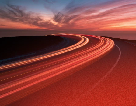 Curved road at dusk with streaks of light trails from vehicles under a colorful sky with clouds.