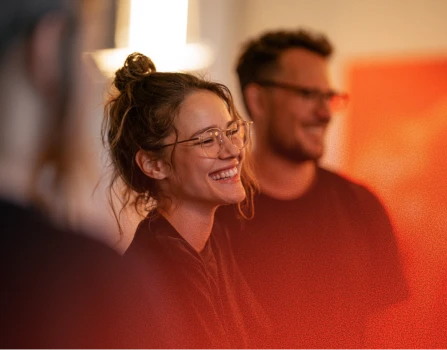 Smiling young woman with glasses and curly hair tied up, sitting indoors with another person in the background, both appearing happy and engaged.