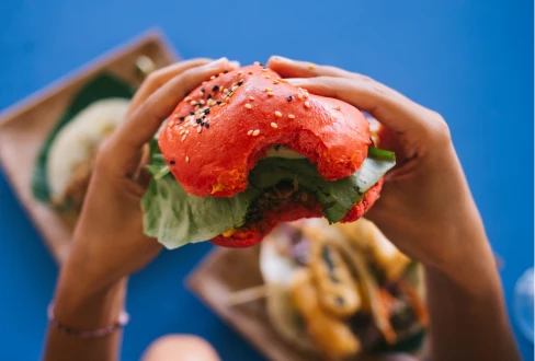 Hands holding a sesame-seed bagel sandwich filled with leafy greens, with another sandwich blurred in the background.