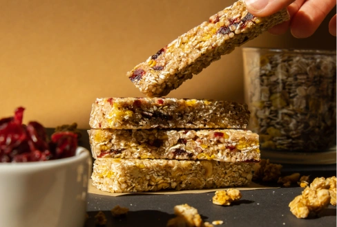 Hand picking up a homemade oat and dried-fruit granola bar from a stack, with dried fruit and seeds in the background.