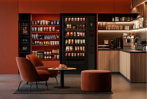 Modern self-service lounge with snack and drink vending machines, coffee station, and rust-colored seating.