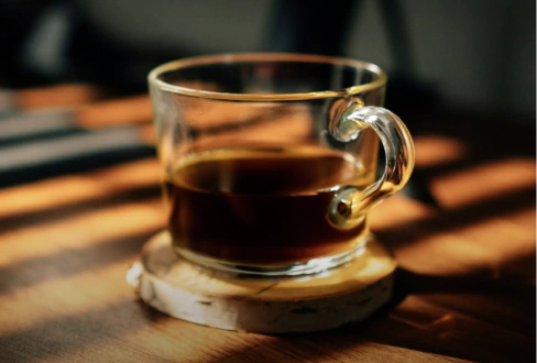 Glass cup of black coffee resting on a coaster on a sunlit wooden table.