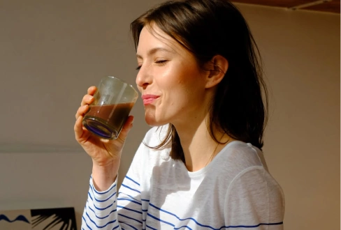 Woman smiling as she takes a sip of a drink from a glass in warm sunlight.