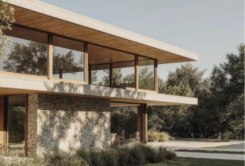 Contemporary house with large windows, stone walls, and a wide overhanging roof, surrounded by trees and greenery.