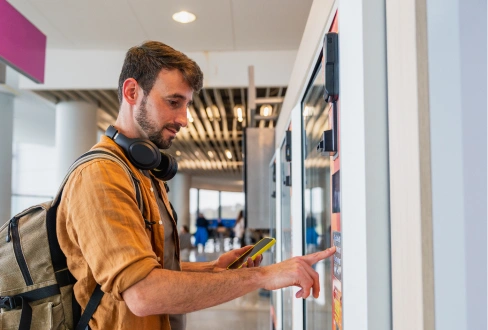 Man with headphones and a backpack using the touchscreen of a vending machine in a modern indoor space.