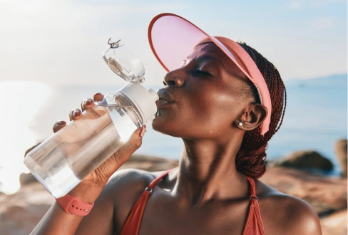 Woman in a pink visor drinking water from a reusable bottle outdoors near the coast.
