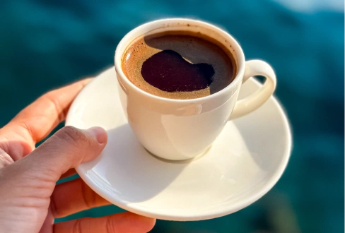 Hand holding a white cup and saucer filled with freshly brewed black coffee.