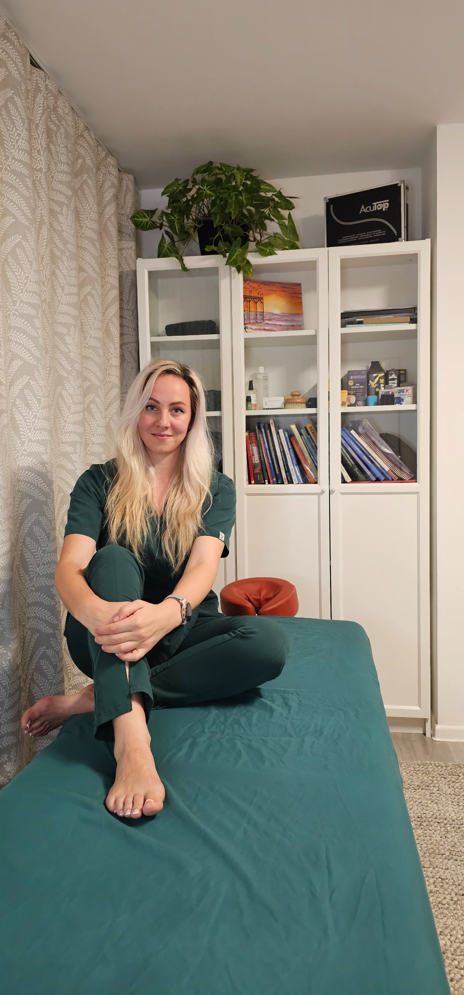 Martyna Nadolska in green scrubs, sitting on the treatment couch in her treatment room