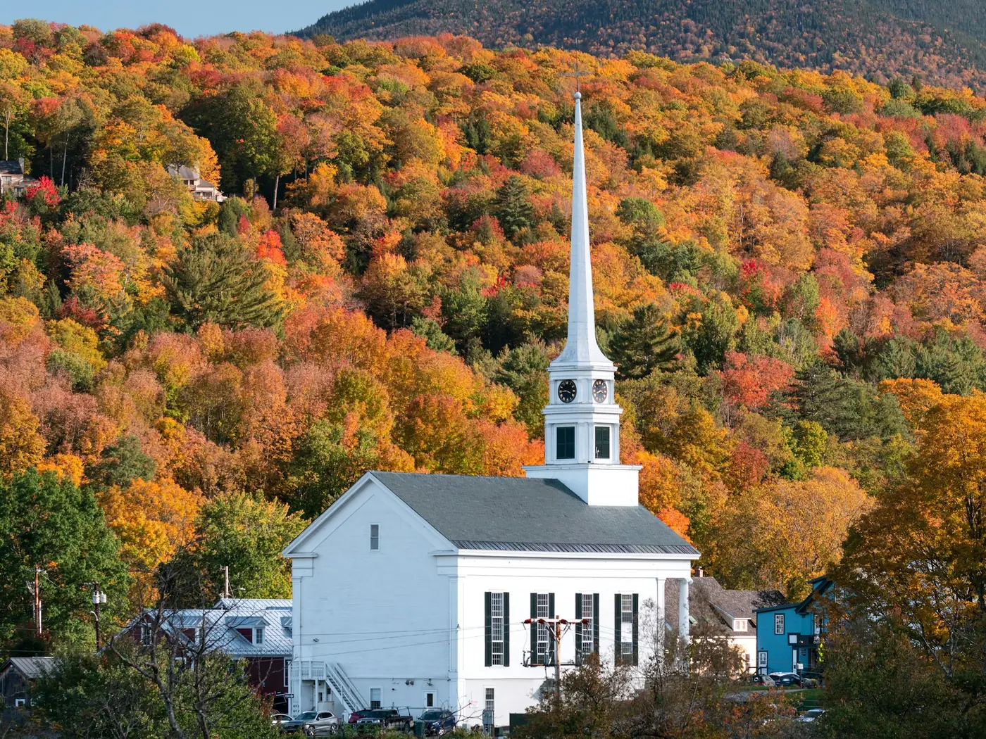 White church with a tall steeple in front of a hillside of autumn trees