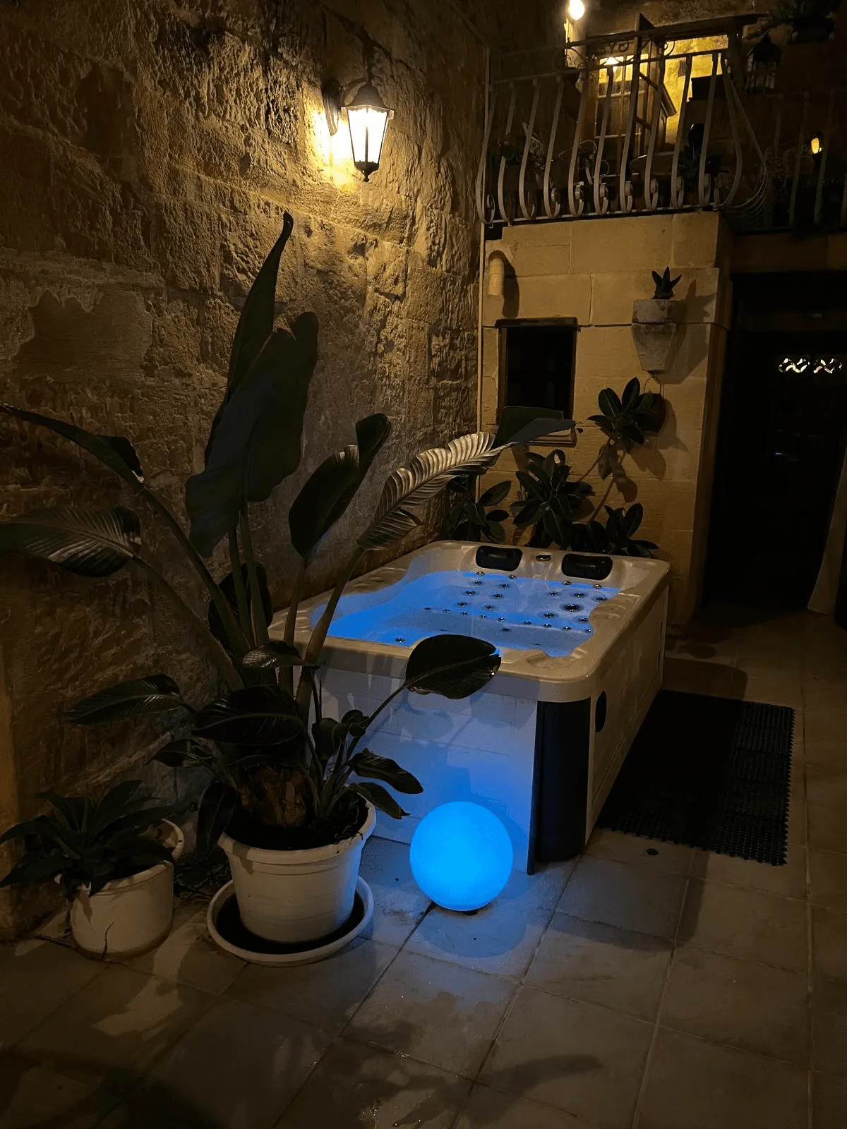 Lit outdoor hot tub at night with plants and a glowing blue orb beside it on stone floor.