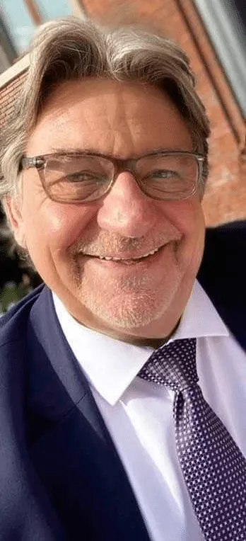 Smiling older man with glasses in a suit and patterned tie against a brick background.