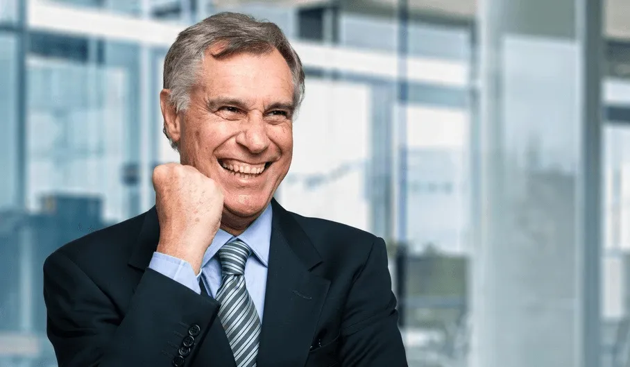Smiling mature man in suit with clenched fist expressing excitement in a modern office setting.
