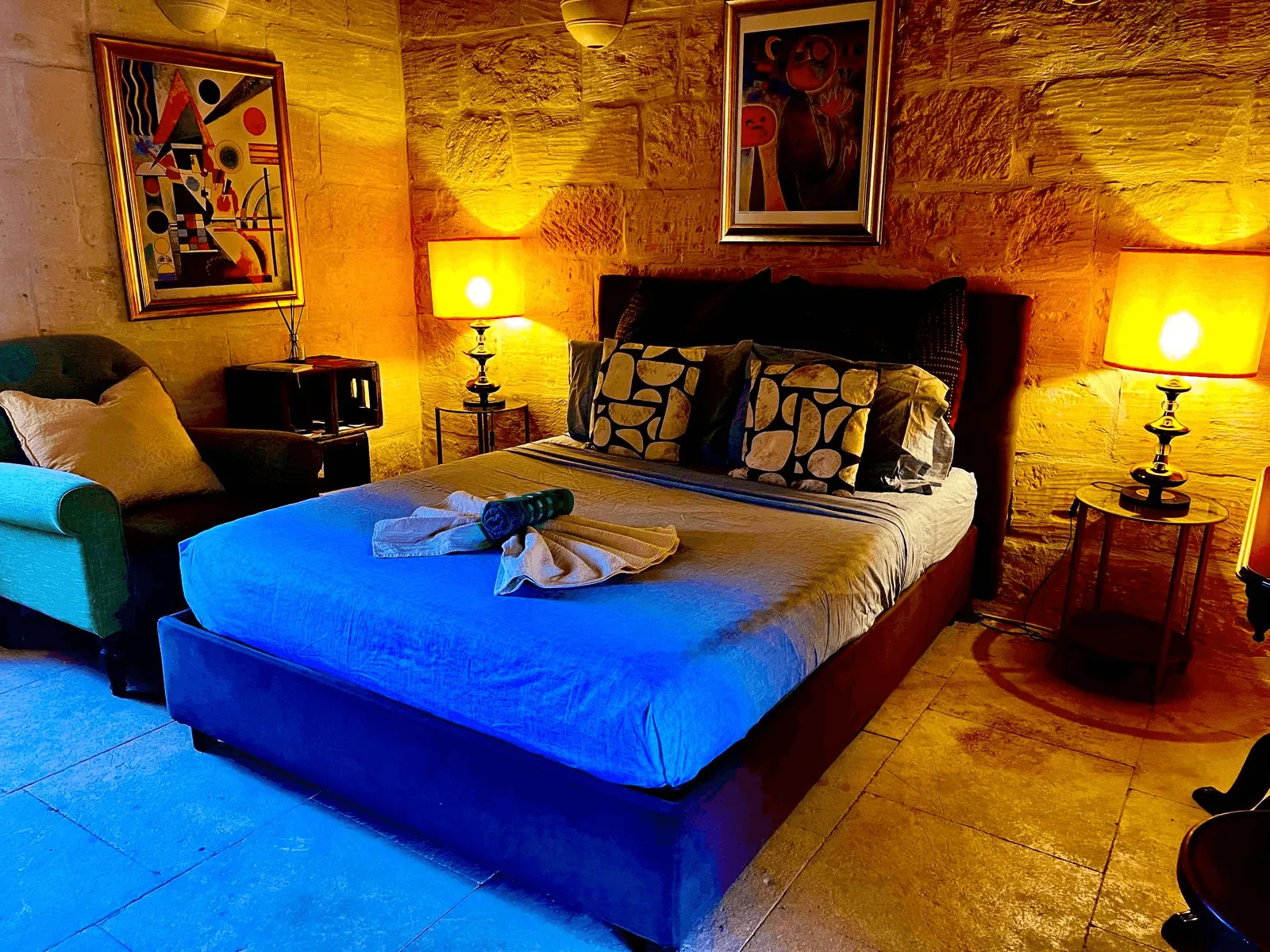 Cozy bedroom with blue bed, patterned pillows, two lamps, armchair, and abstract paintings on stone walls.