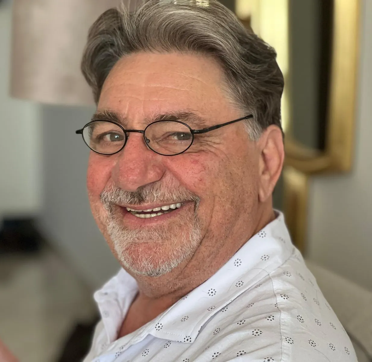 Smiling older man with gray hair, beard, glasses, wearing a white patterned shirt indoors.
