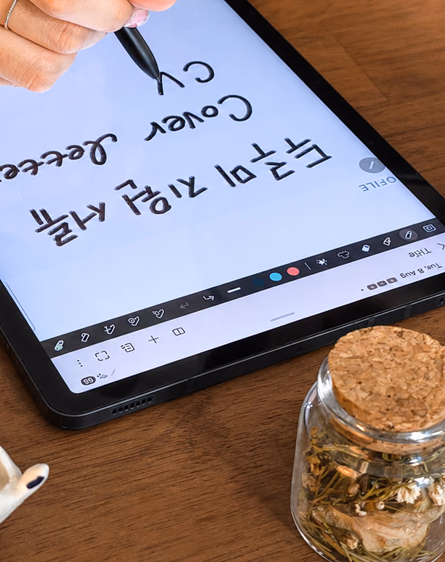 Hand writing on a tablet screen with a black stylus on a wooden desk with a jar of dried herbs.