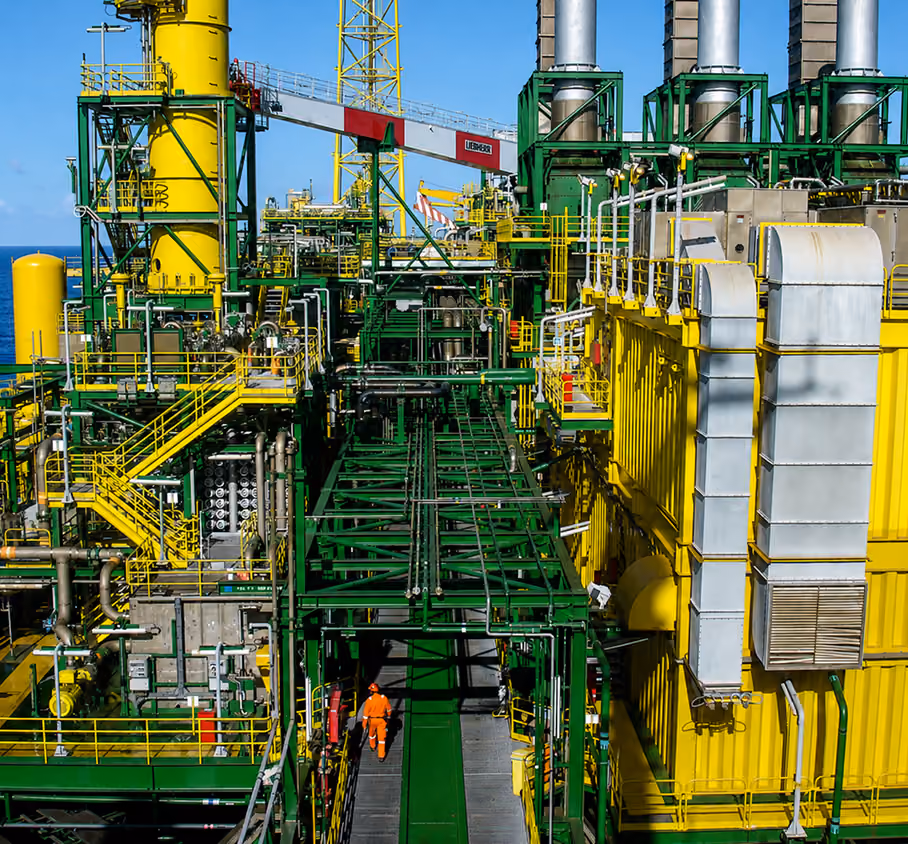Industrial oil rig platform with yellow and green structures and a worker in orange safety gear walking.