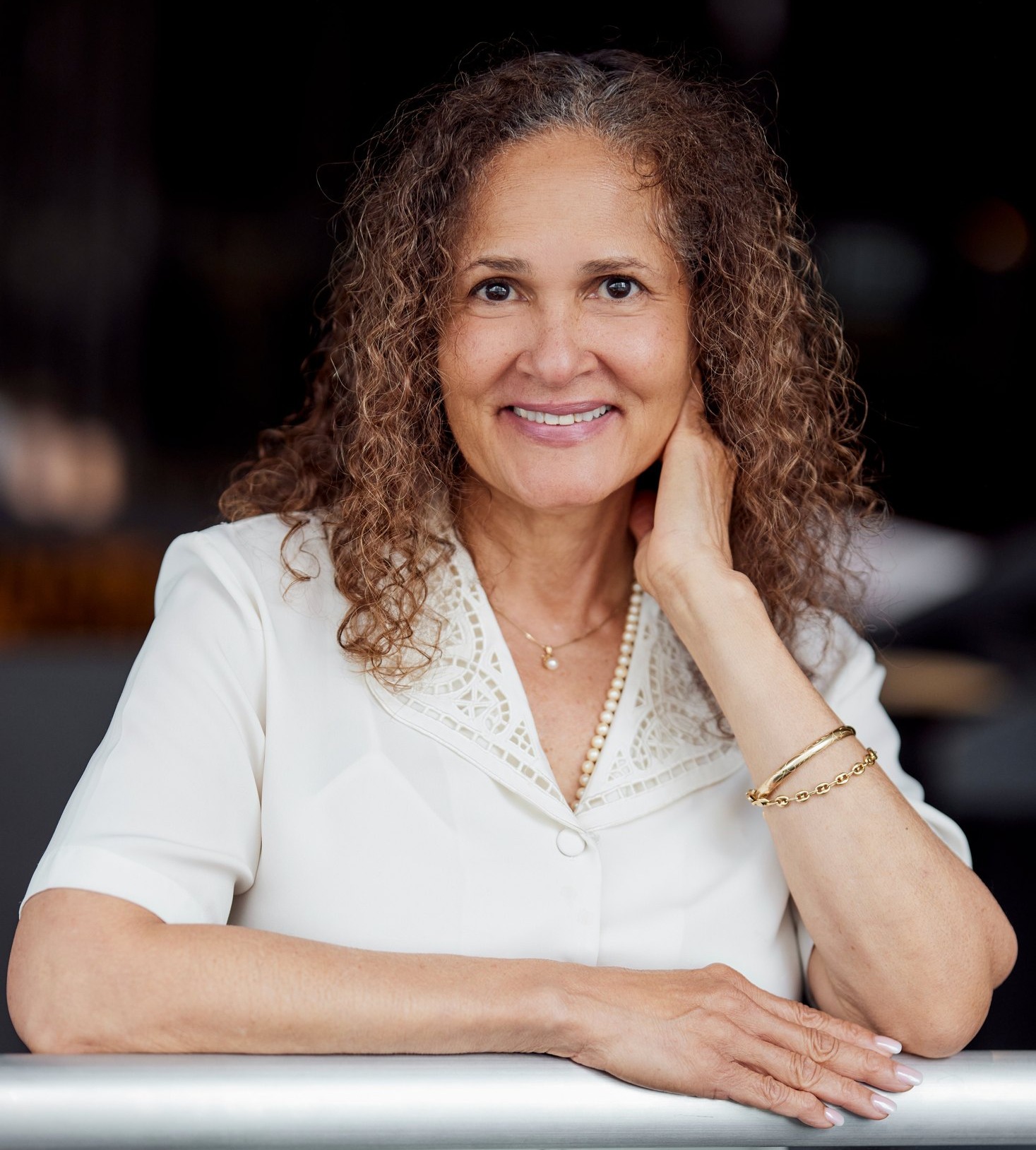 Portrait of Elizabeth Lawton, smiling, in a cream jacket, leaning on a railing against a dark background.
