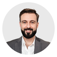 Smiling man with dark hair, beard, and mustache wearing a gray suit and white shirt against a light background.