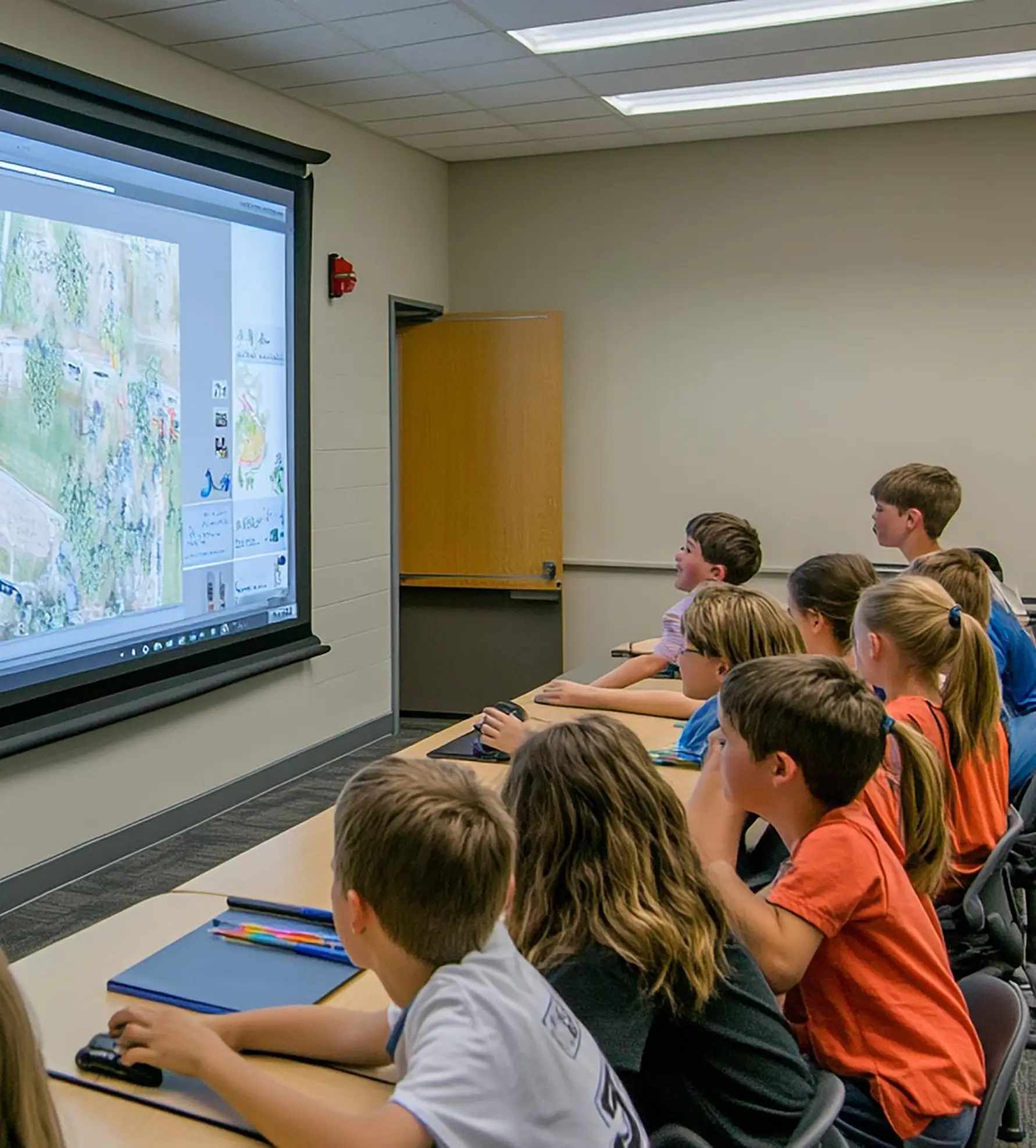 Children seated at desks attentively watching a projected map on a classroom screen.