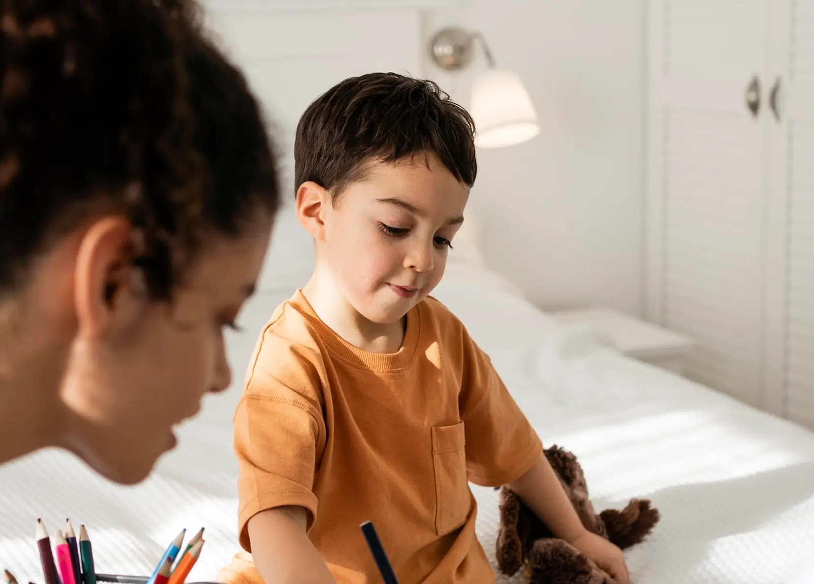 Young boy in an orange shirt drawing on a bed with colored pencils and a woman watching him.