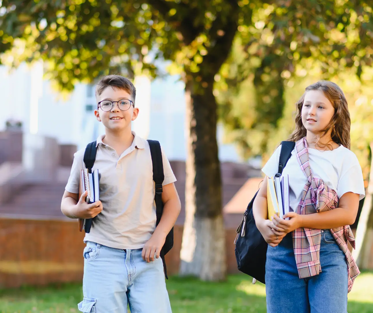 Two schoolchildren with backpacks and books walking outdoors under trees on a sunny day.