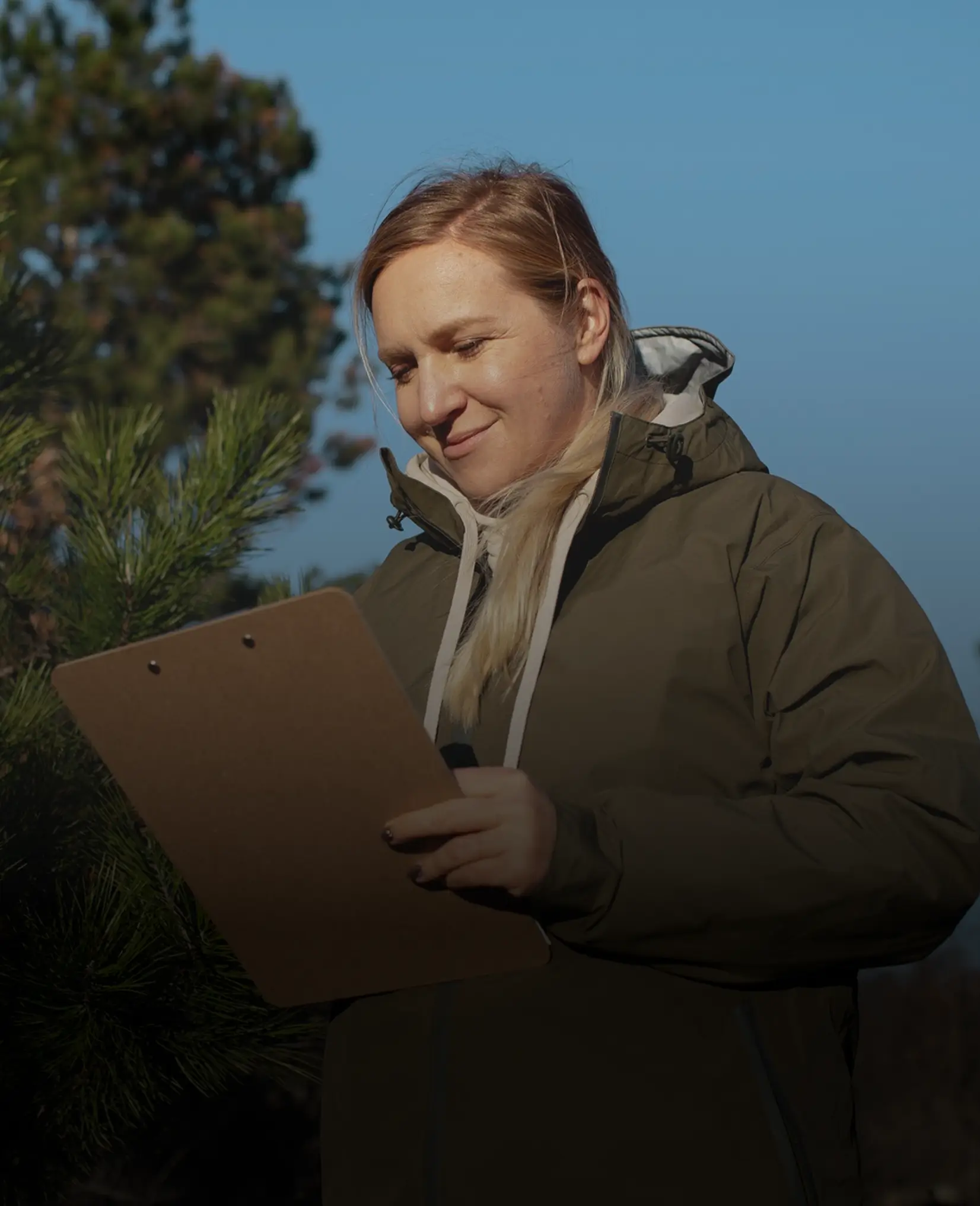 Woman in green jacket outdoors holding a clipboard, smiling while looking at it near pine trees.