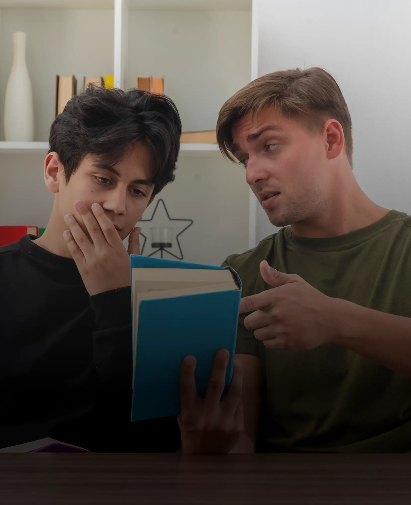 Two young men sit together with a book, one looking concerned and the other explaining.