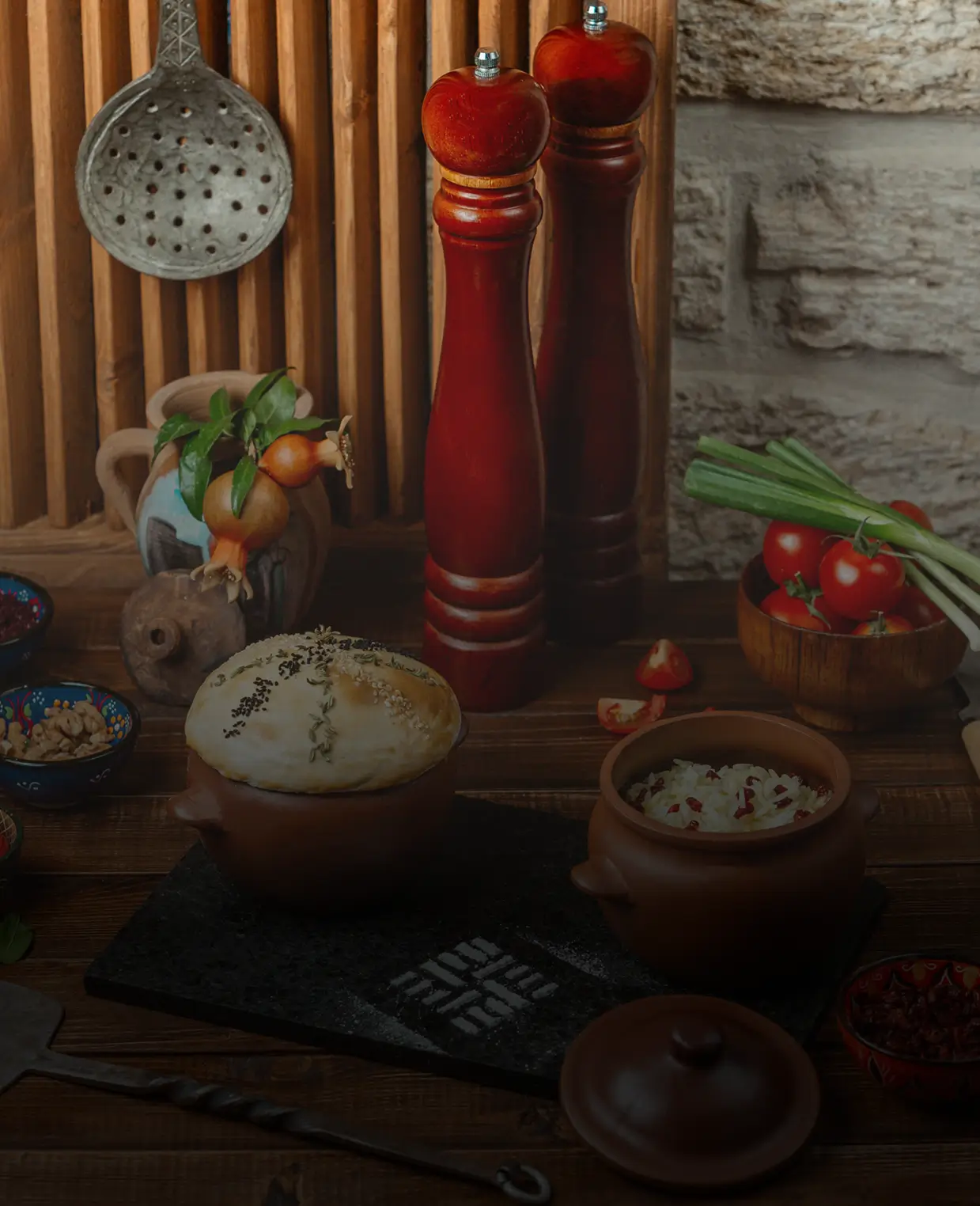 Two brown pots with food, wooden pepper mills, fresh tomatoes, and green onions on a rustic wooden table.