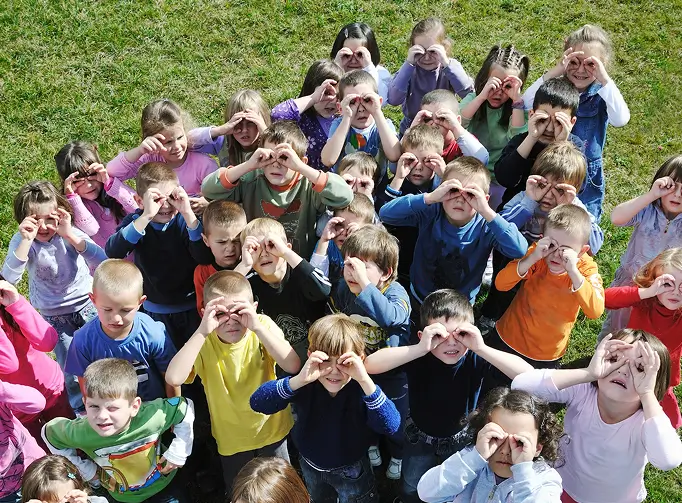 Group of preschool kids outdoors making binocular shapes with their hands over their eyes on grass.