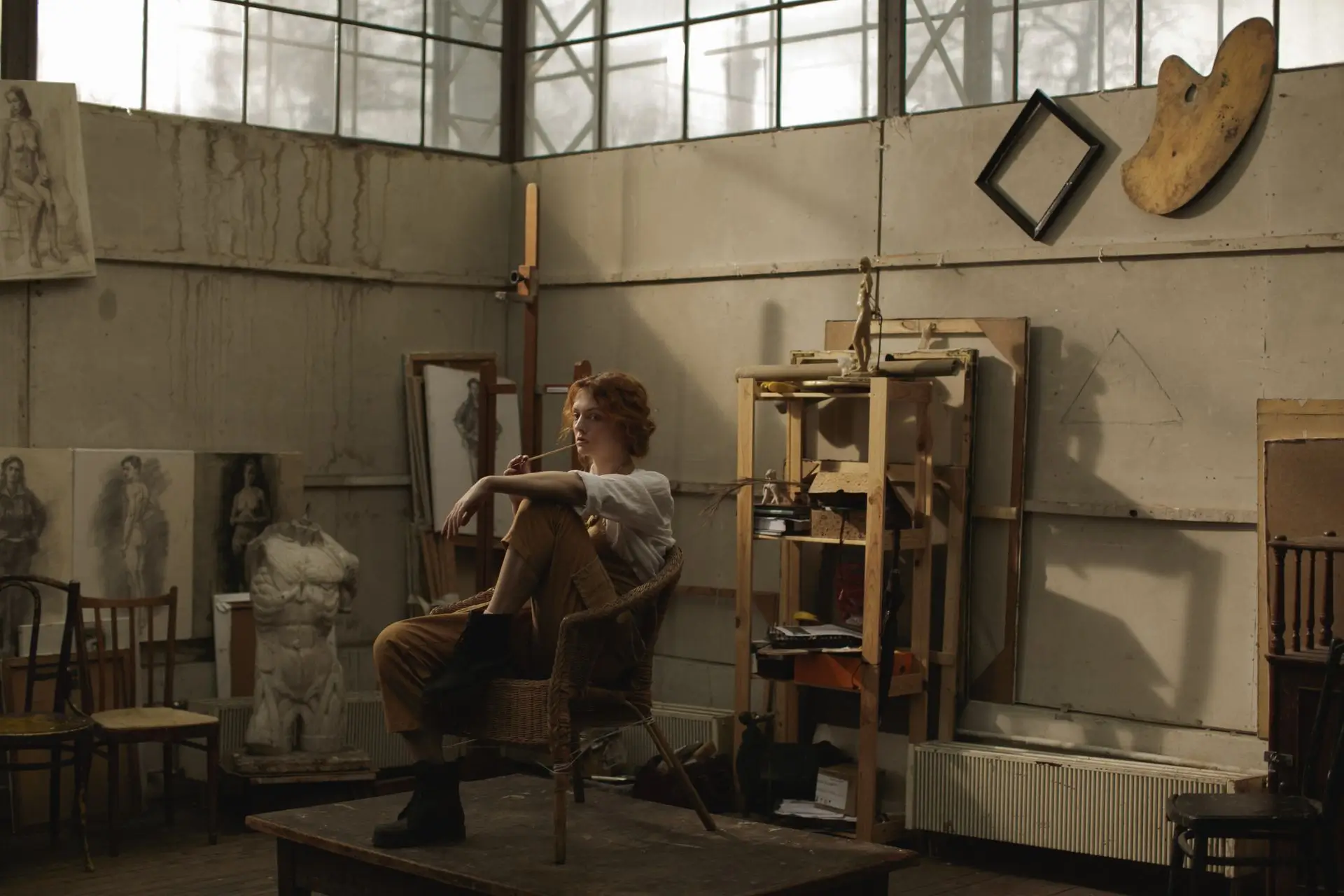 Red-haired artist sitting on a wicker chair in a studio surrounded by sketches and sculptures.