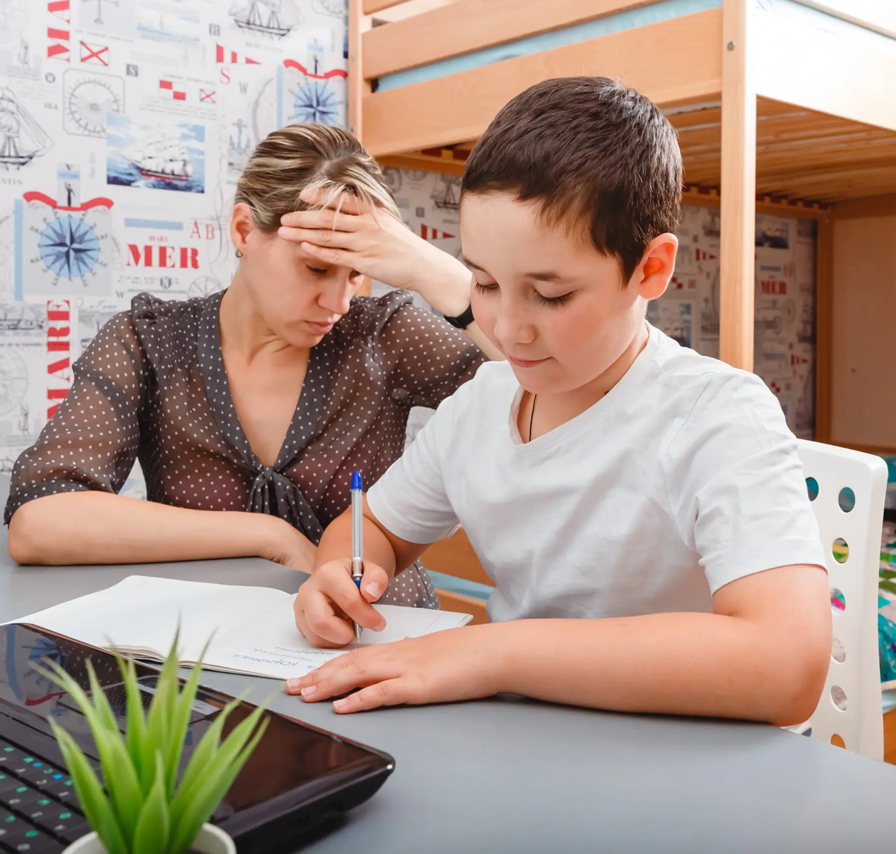 Boy in white shirt writing in notebook while a stressed woman watches him at a table.