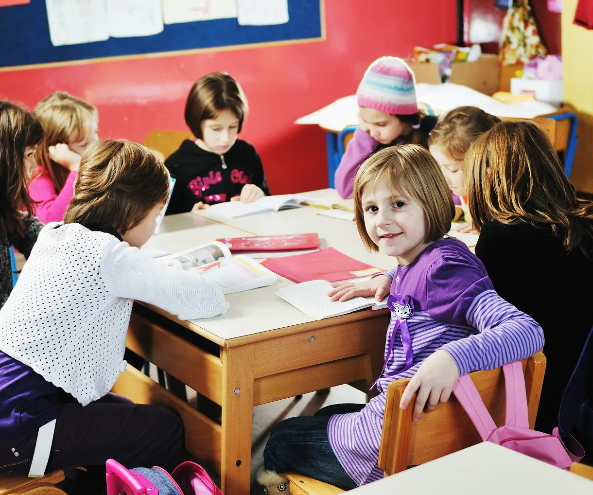 Group of young children sitting around a table in a classroom, reading books and studying, with one girl in a purple striped shirt smiling at the camera.
