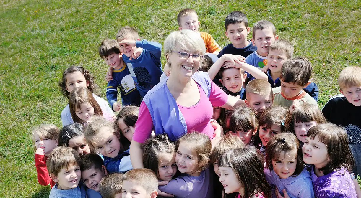 A smiling female teacher with short blonde hair and glasses surrounded closely by a diverse group of young children on a grassy field, all looking happy and engaged.