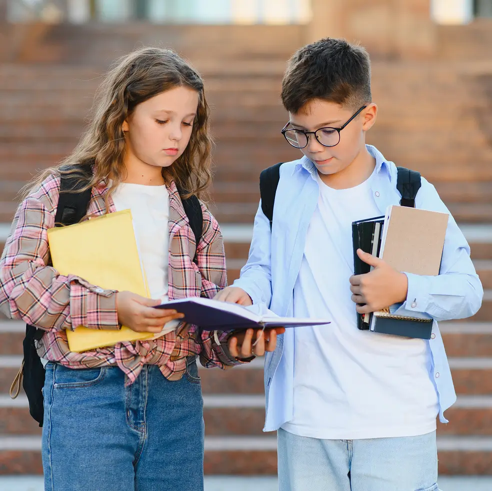 Two schoolchildren standing outdoors in front of stairs, a girl holding a yellow folder and a boy with glasses holding several notebooks, both looking at an open book the girl is holding.