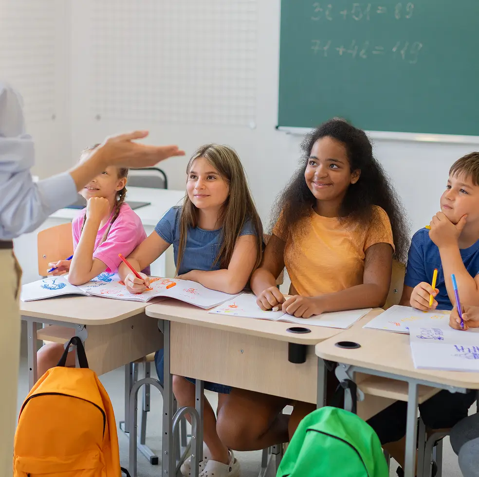 Four children sitting at desks in a classroom, engaged and smiling while a teacher speaks and gestures with their hand in front of them, with notebooks and colored pens on the desks and backpacks hanging nearby.