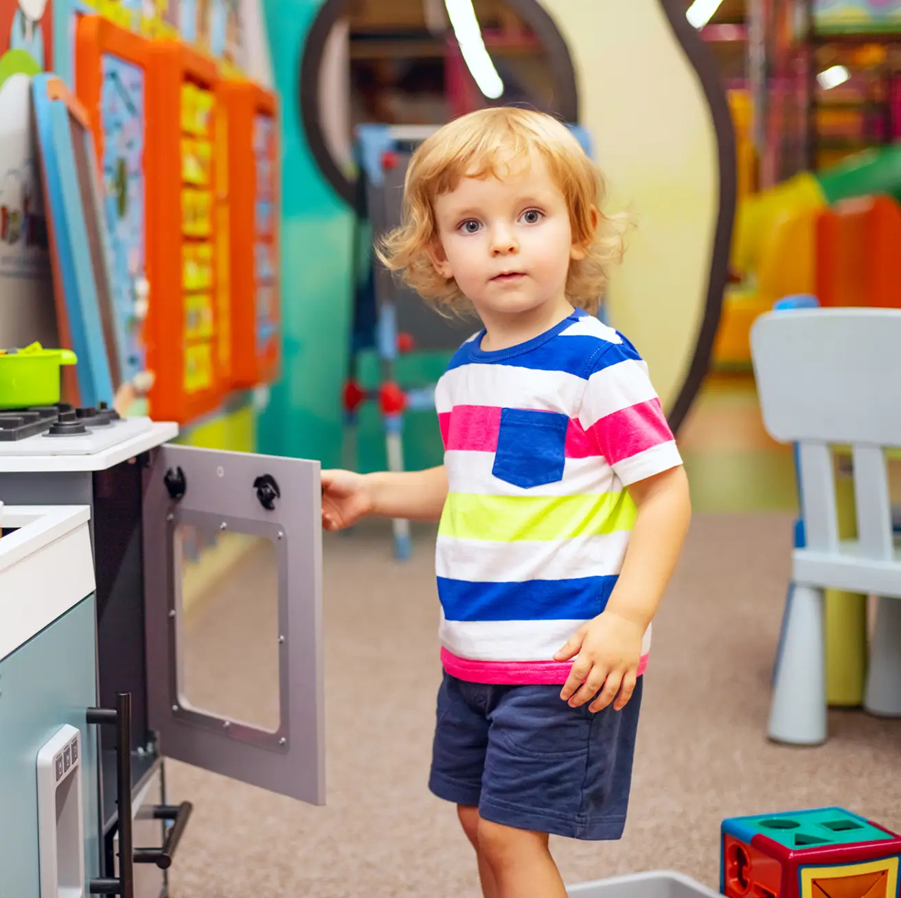 Young child with blonde curly hair wearing a colorful striped shirt and blue shorts playing with a toy oven in a vibrant indoor playroom.