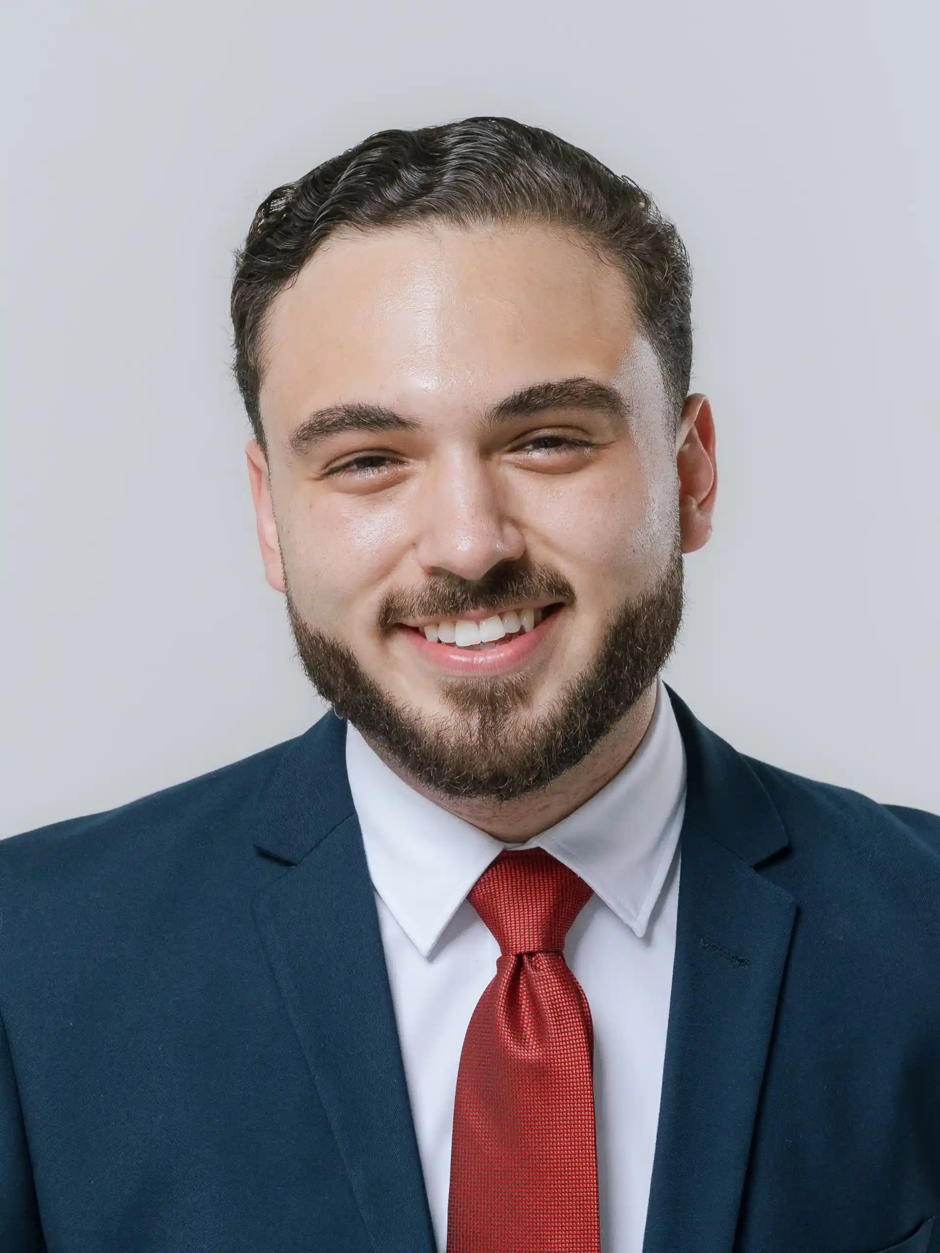 Smiling young man with neatly groomed dark hair and beard, wearing a dark blue suit, white shirt, and red tie, against a light gray background.