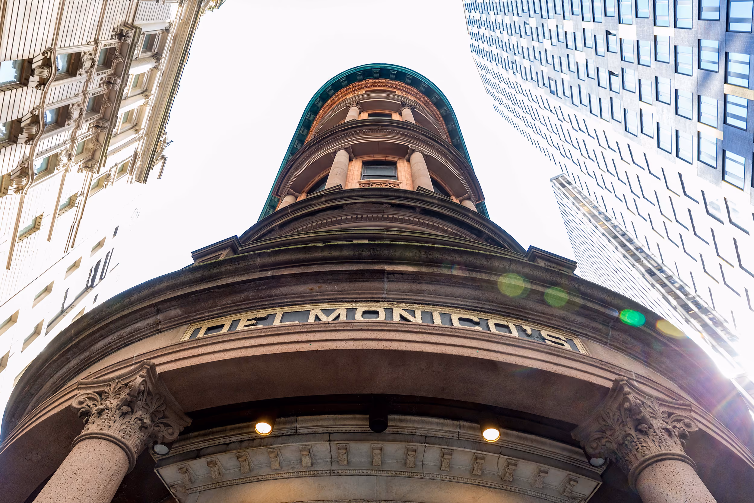 Low-angle view of the Delmonico's building with ornate columns and tall modern buildings around.