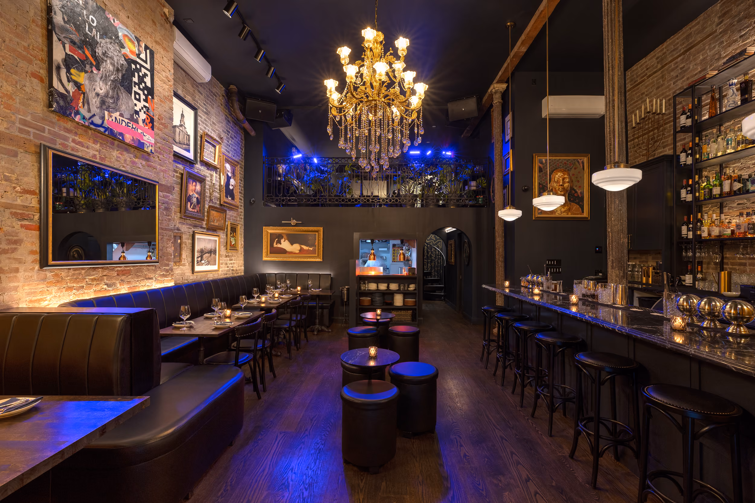Cozy bar interior with leather seating, framed art on exposed brick walls, chandelier, and marble countertop.