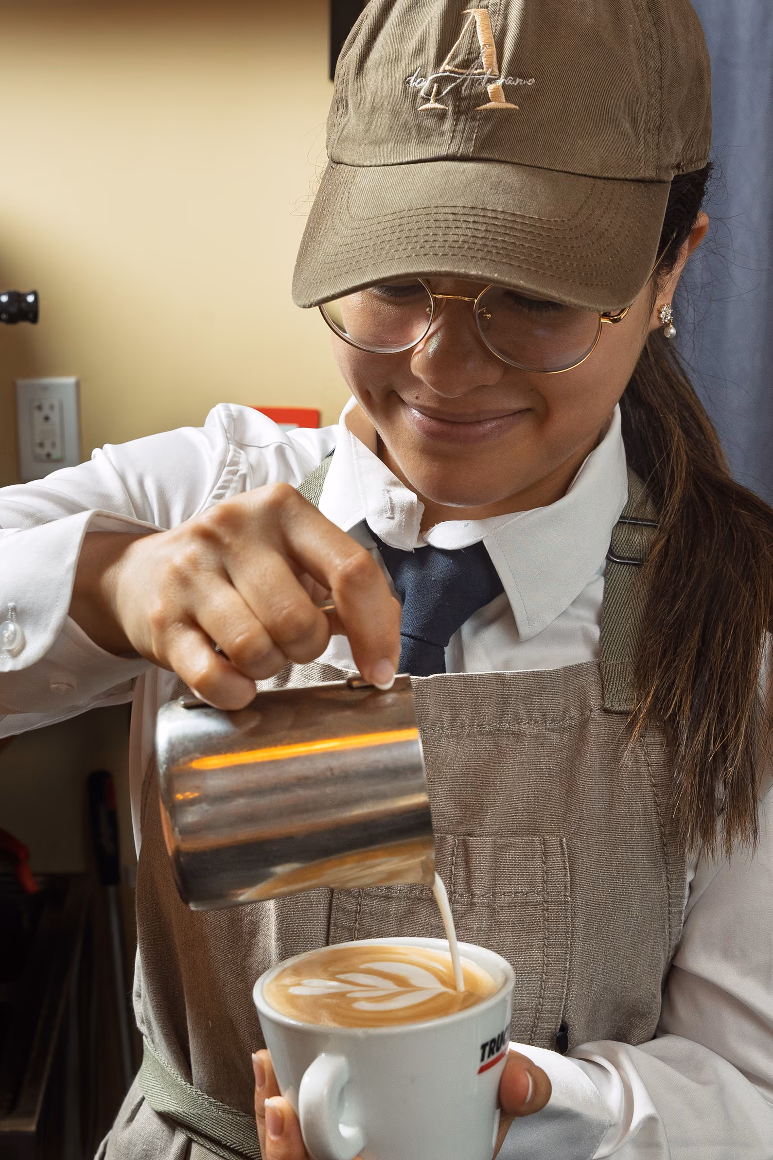 Barista in glasses and hat pouring milk foam to create latte art in a white coffee cup.
