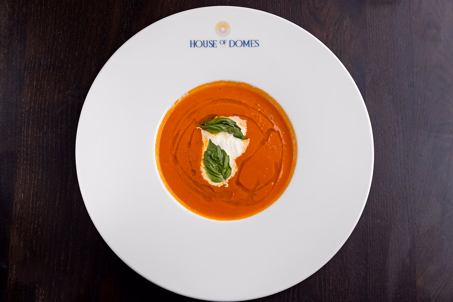 White plate with logo, holding creamy tomato soup garnished with cream and basil leaves.