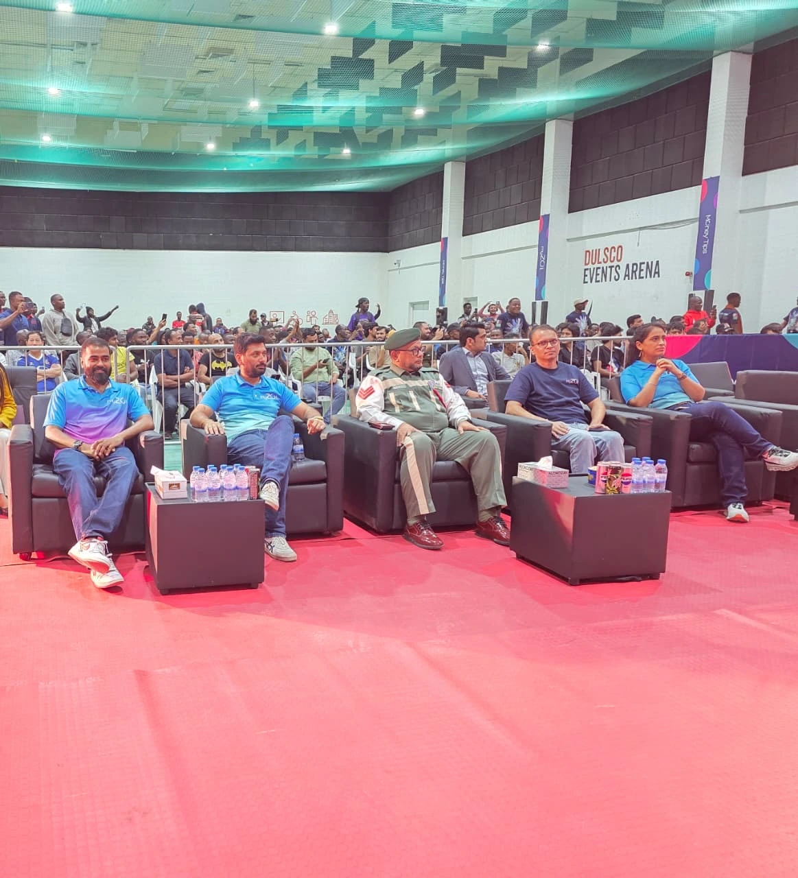Guests in blue shirts seated in armchairs at the front of a community event at Dulsco Events Arena
