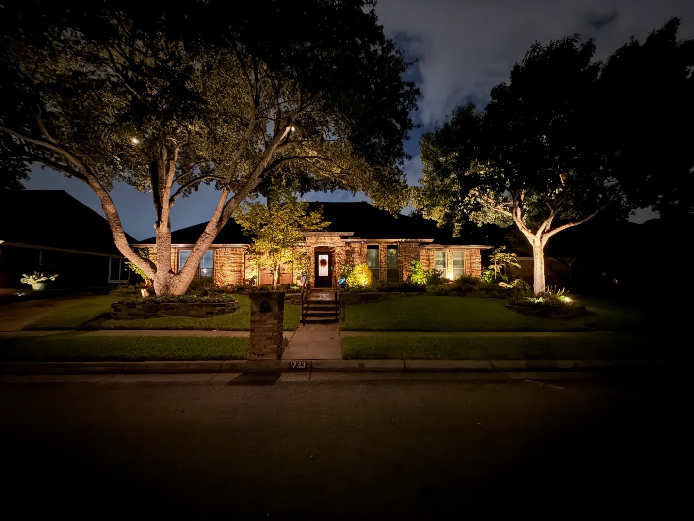Front-yard landscape lighting illuminating a brick home and mature trees at night
