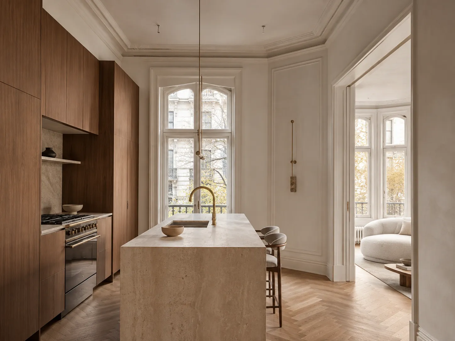 Haussmannian-style kitchen with herringbone parquet floors, walnut cabinetry, brass fixtures, and an arched window opening onto a curved boucle sofa in the adjoining living room.