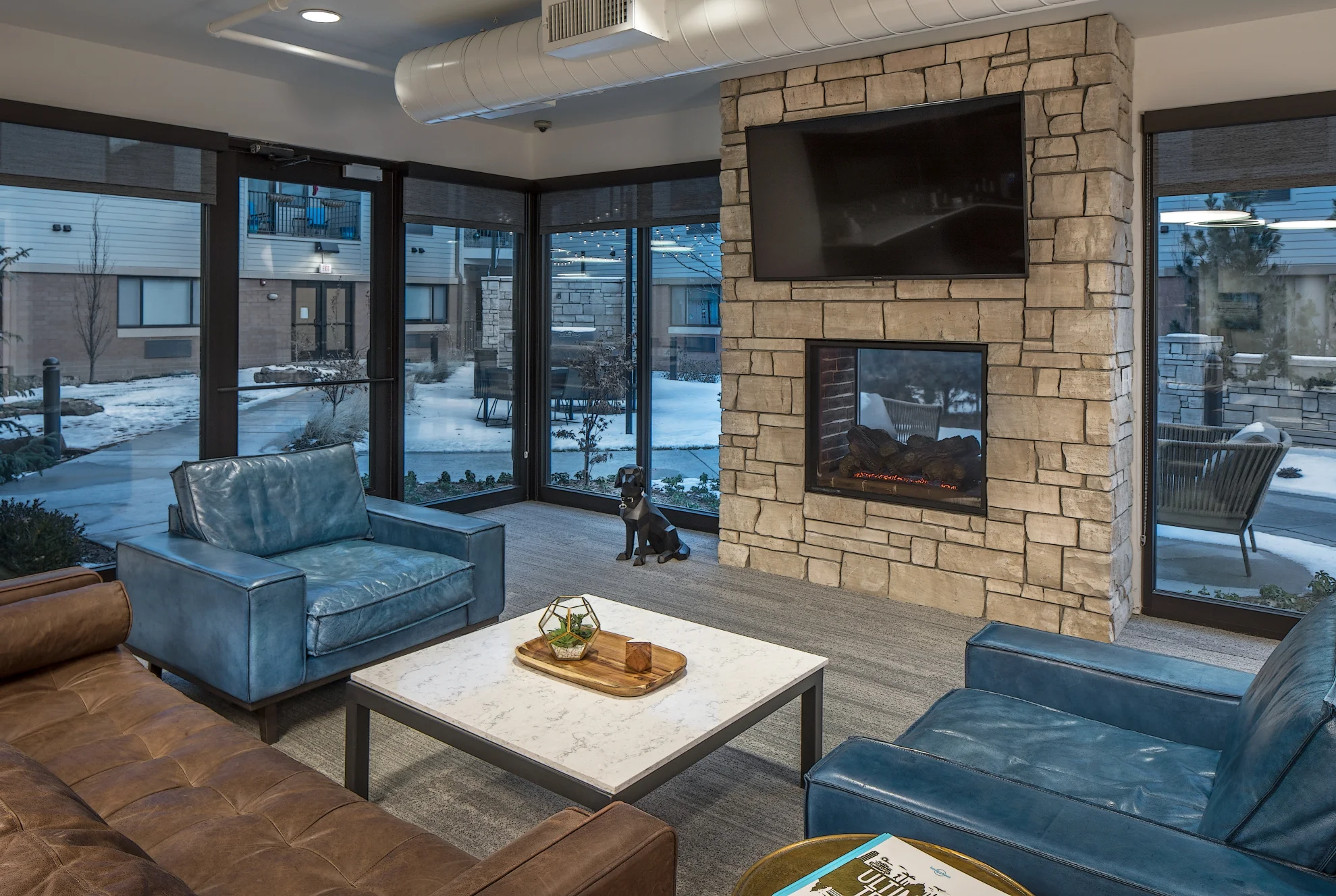 Lounge with teal armchairs and sofas around a coffee table beside a stone fireplace and floor-to-ceiling windows