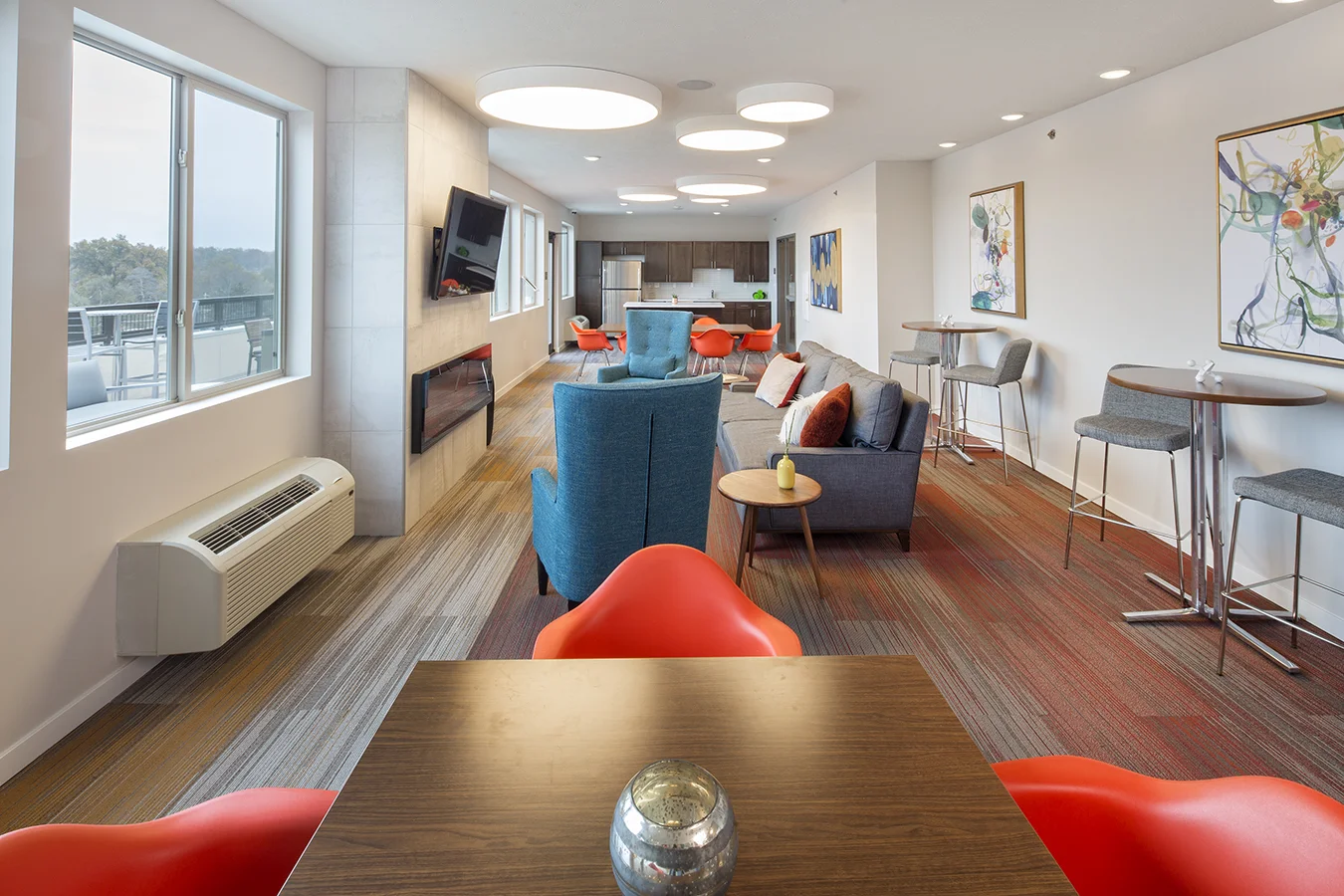 Square at 48 community room in Lincoln with orange dining chairs, teal lounge seating and large windows
