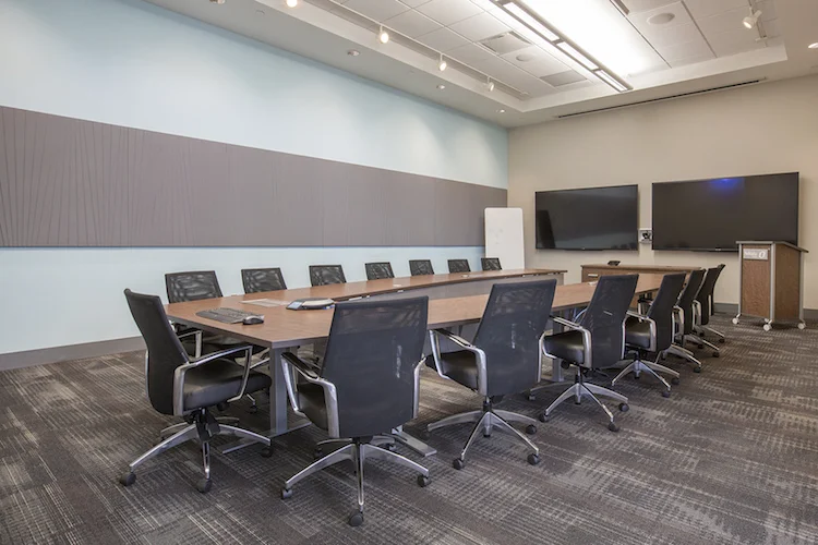 Boardroom with a long wood conference table, black leather chairs and dual display screens
