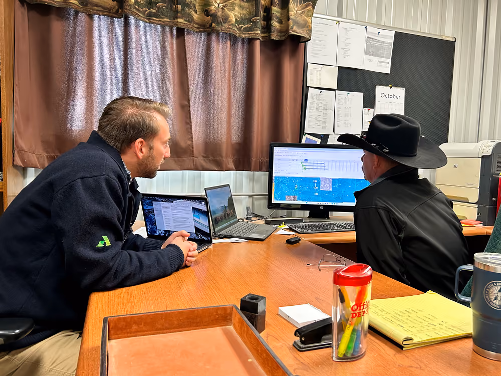 Two men sit at an office desk focused on a desktop monitor displaying a map and spreadsheet; one wears a black cowboy hat and jacket, the other a dark fleece jacket, with laptops, office supplies, and notes on the table.