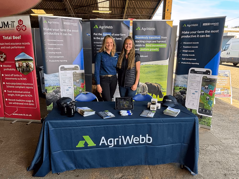 Two women standing behind an AgriWebb promotional table at an indoor event, with banners highlighting farm productivity, crop and herd management software, and product features like movement and treatment records.