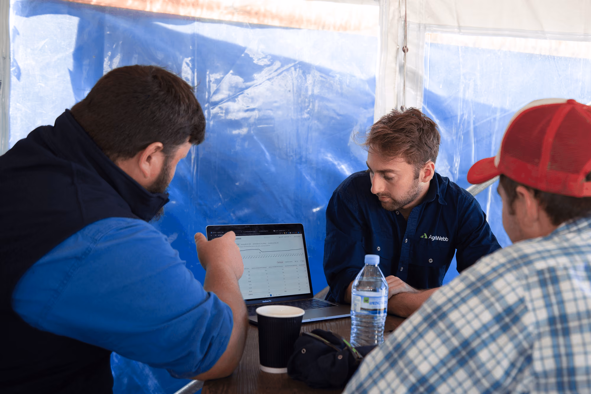 Three men sitting at a table inside a tent, focused on a laptop screen displaying data; one man wears a navy shirt with 'AgriWebb' logo, another wears a blue shirt with a black vest, and the third wears a red cap and checkered shirt. There is a water bottle and two cups on the table.